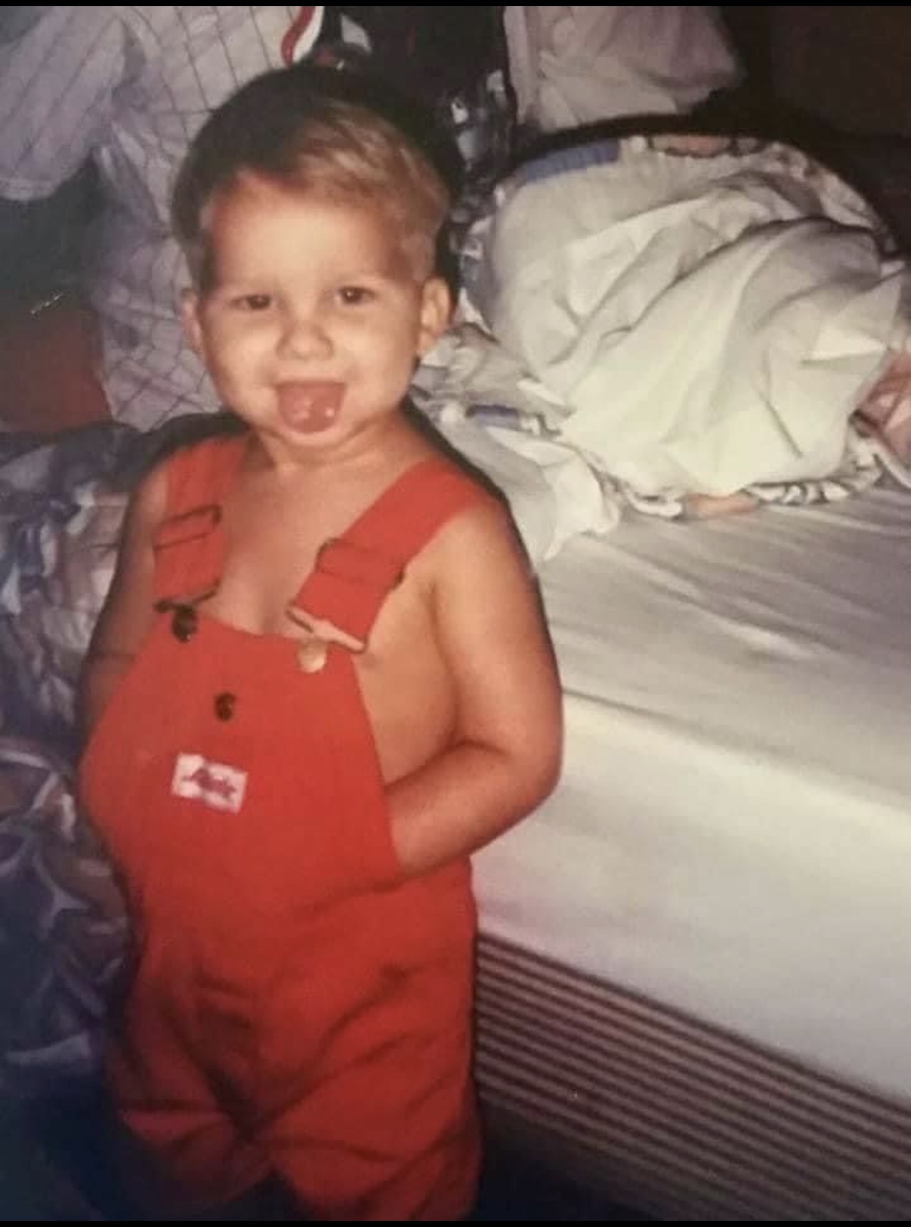 A young Dustin wearing red overalls, grinning with his tongue out.